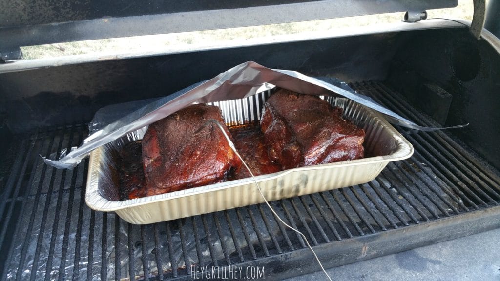Two pork butts in an aluminum pan with a meat thermometer in one of the pork butts, on the grill.