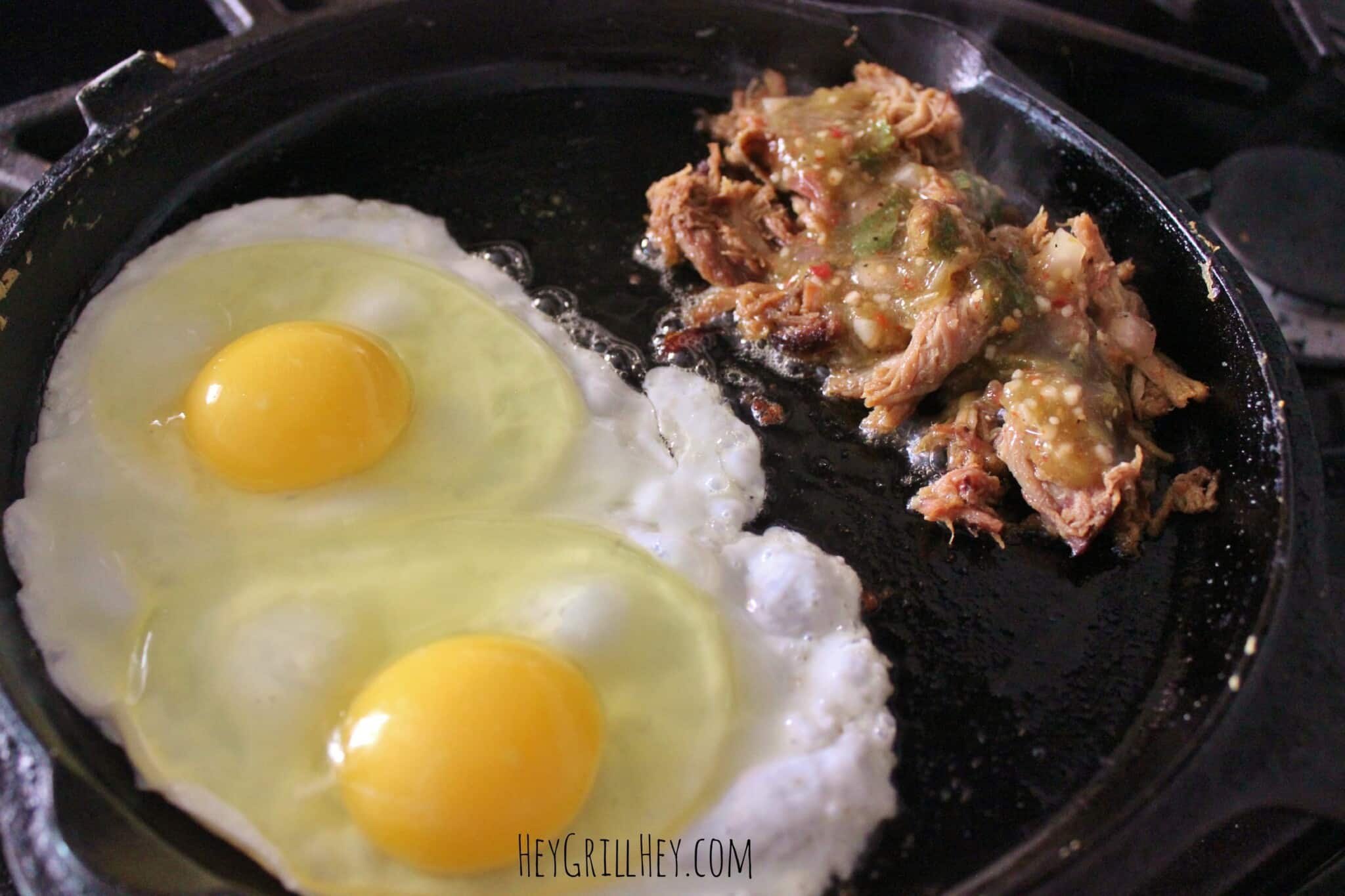 Hot skillet topped with two eggs cooking and a pile of pulled pork topped with green salsa.