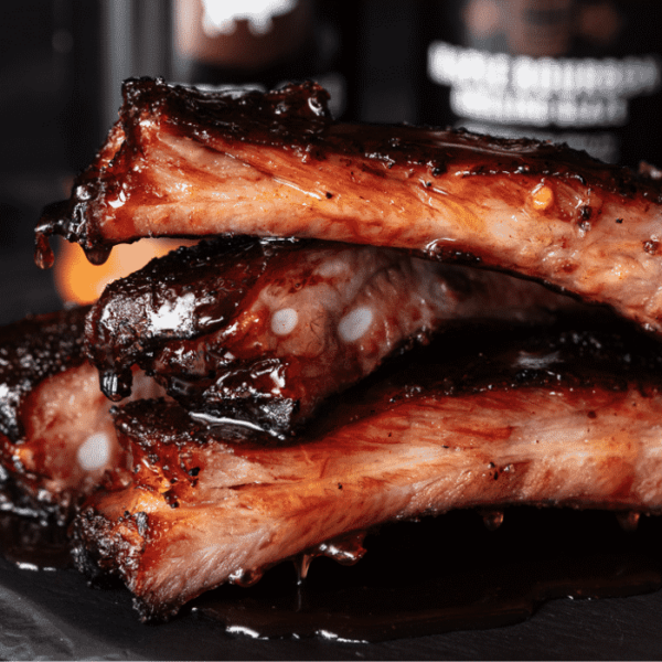 Sauced and smoked ribs stacked in front of bottles of BBQ sauce.