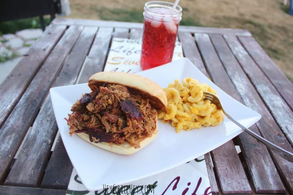 Pulled Pork Sandwich on a bun served on a white plate with a side of macaroni and cheese and a glass of red juice.