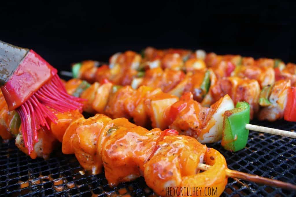 Close up shot of cooked and cubed chicken and peppers on the grill grate being brushed with buffalo Sauce.