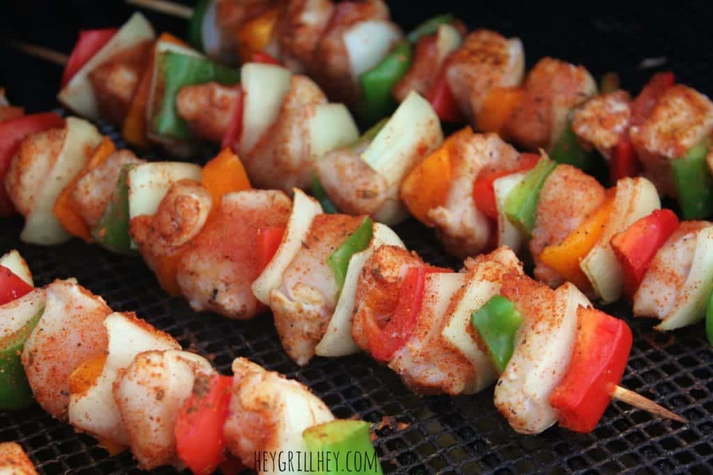 Close up shot of wooden skewers with pieces of seasoned chicken, bell peppers, and onions, lying directly on the grill grate.