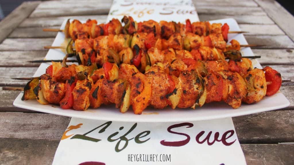Finished Grilled Buffalo Chicken skewers presented on a white plate, which is on top of a decorative dish towel that reads "Sweet Life". All on a wooden table outside.