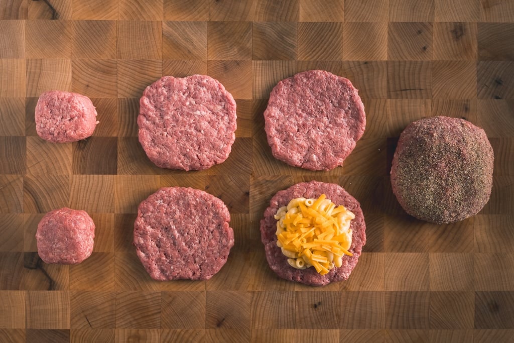 Hamburger patties arranged on a cutting board in various states of being stuffed with mac and cheese.