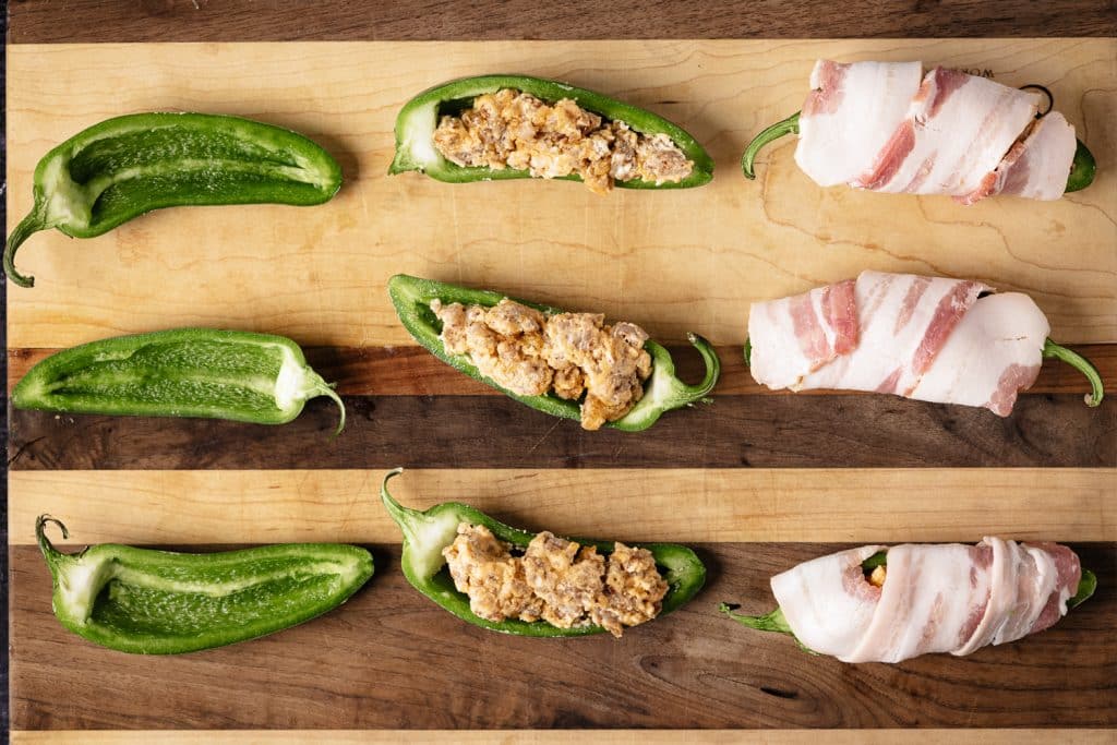 Halved jalapenos on a wooden cutting board in various stages of being stuffed and wrapped in bacon.
