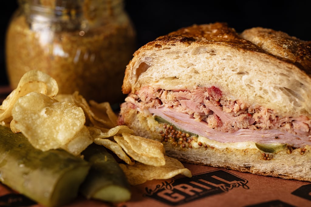 Cuban sandwich on peach butcher paper next to potato chips and a jar of spicy mustard.