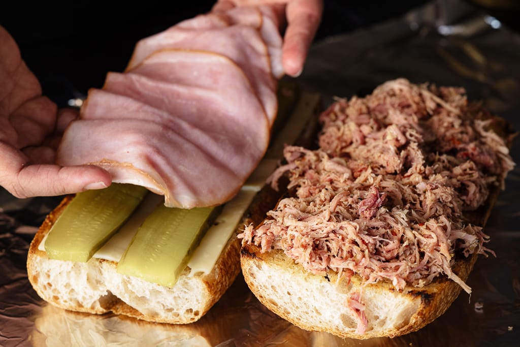 Shredded pork on half a loaf of French break next to the other half topped with sliced pickles and ham.
