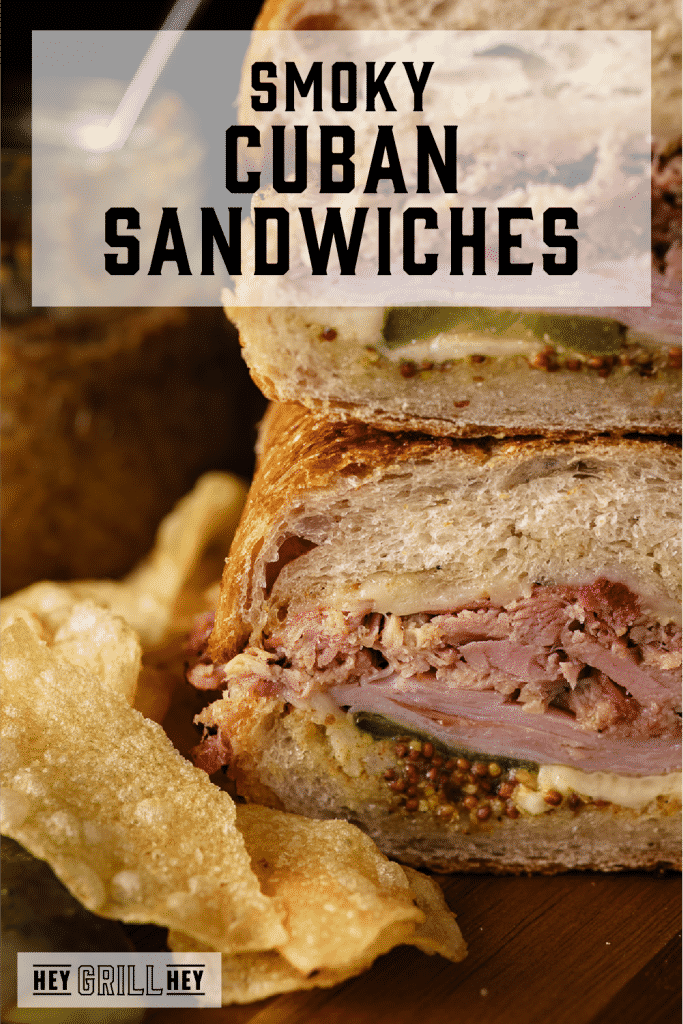 Two Cuban sandwiches stacked on a wooden board next to potato chips with text overlay - Smoky Cuban Sandwiches.