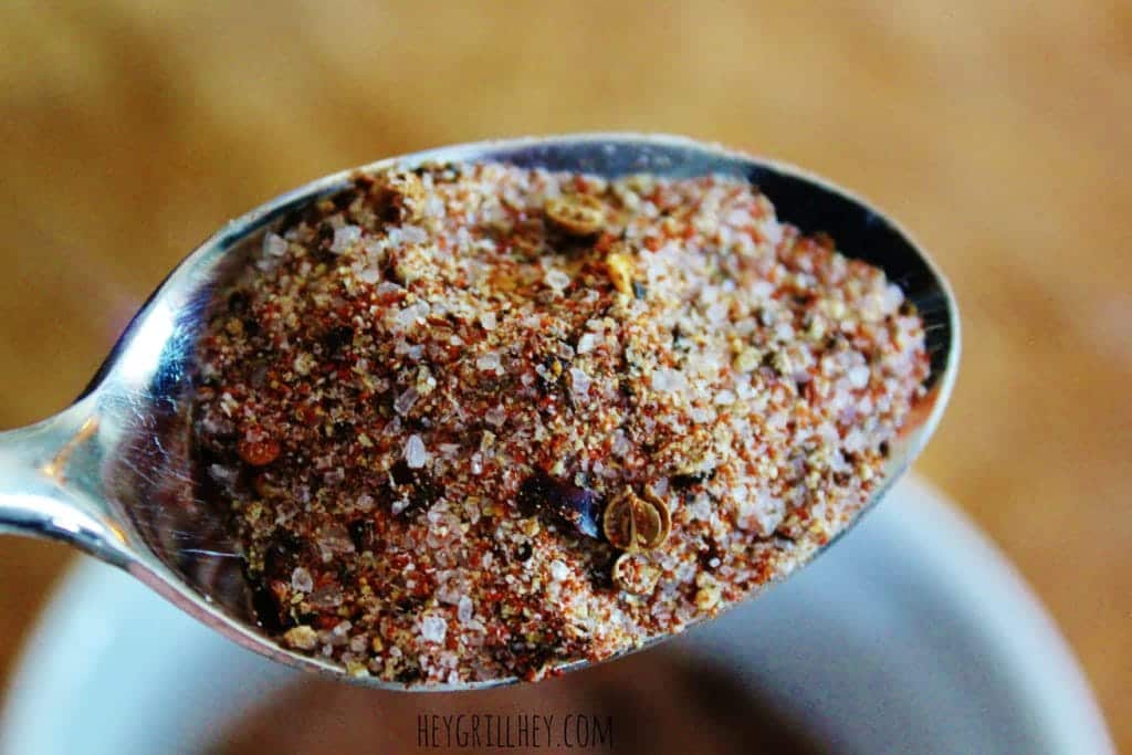 homemade steak rub in a spoon above a white bowl full of more seasoning on a wood cutting board.