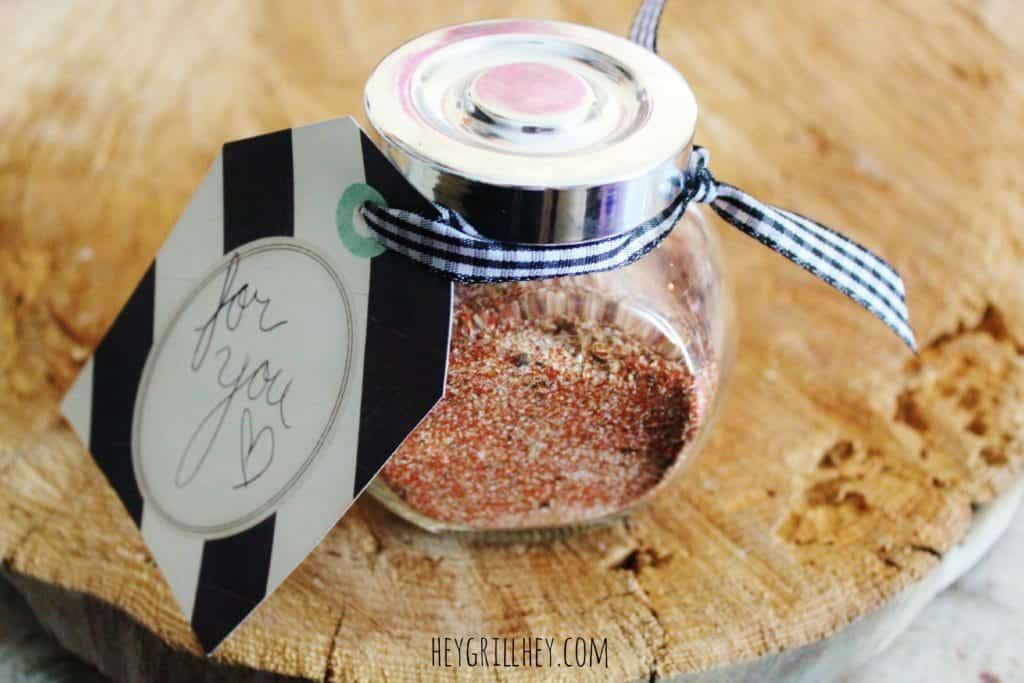 Homemade steak rub in a glass jar with silver lid tied with black and white ribbon and gift tag on a wood cutting board.