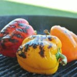 Red, yellow, and orange bell peppers roasting on a charcoal grill.