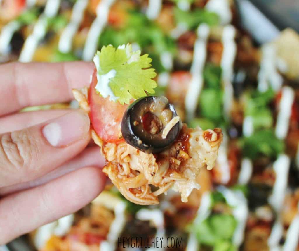 Hand holding a tortilla chip topped with BBQ chicken, cheese, tomato, black olive, and cilantro.