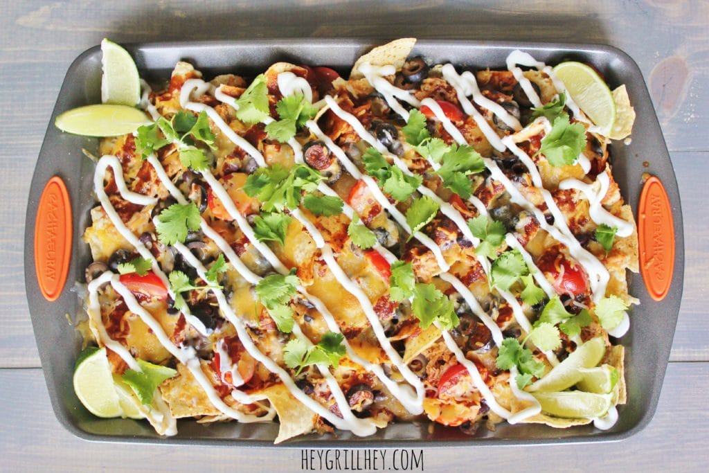 BBQ chicken nachos in a metal baking dish topped with cheese, tomatoes, black olives, sour cream, cilantro, and lime wedges.