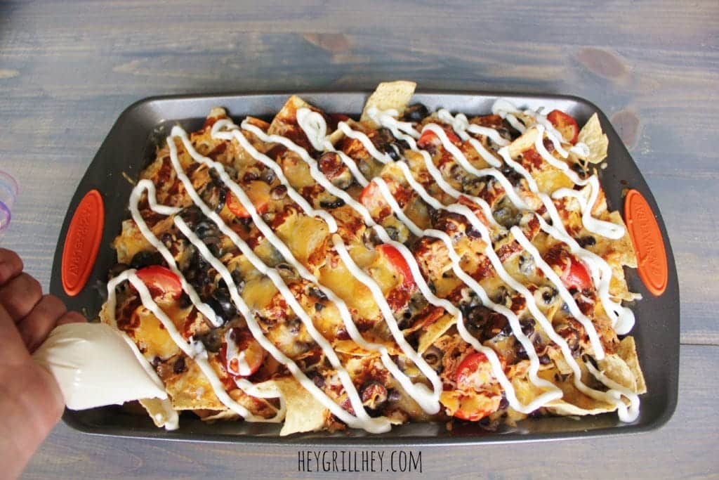Tortilla chips topped with BBQ chicken, tomatoes, black olives, and piped sour cream in a metal baking sheet.