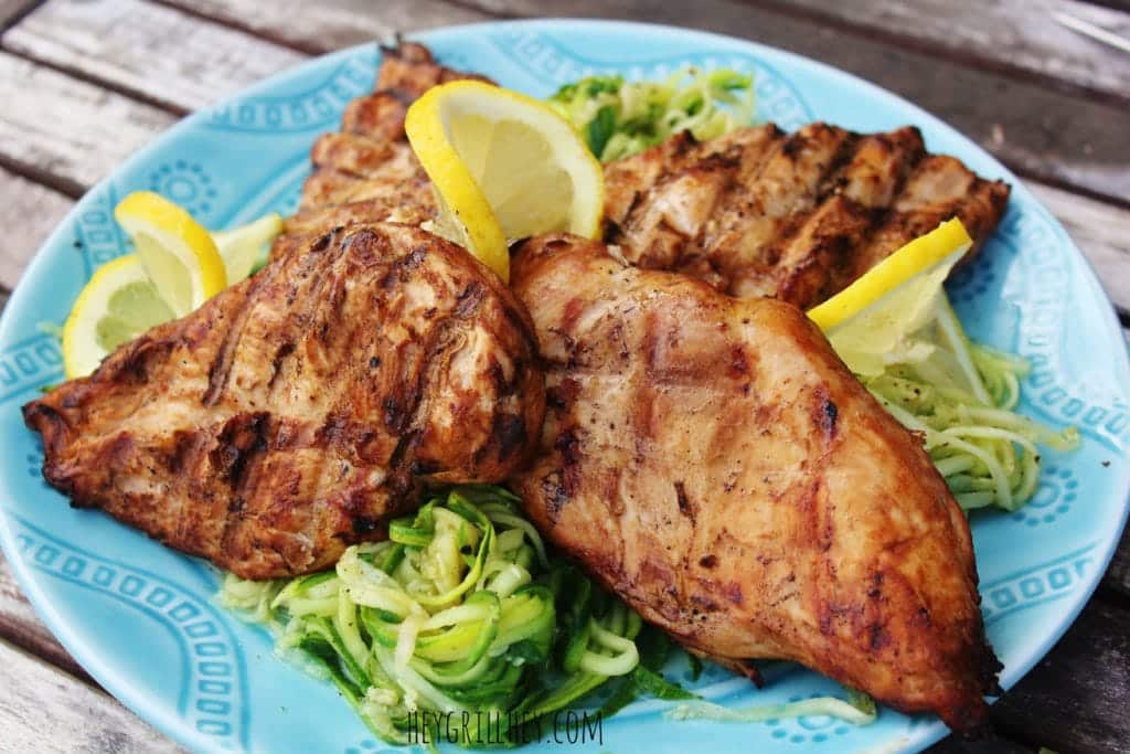 Four grilled chicken breasts on a blue plate with zucchini noodles and lemon slices