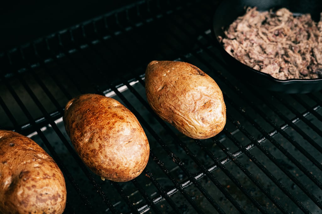 Whole russet potatoes in a BBQ grill next to a cast iron skillet of brisket.