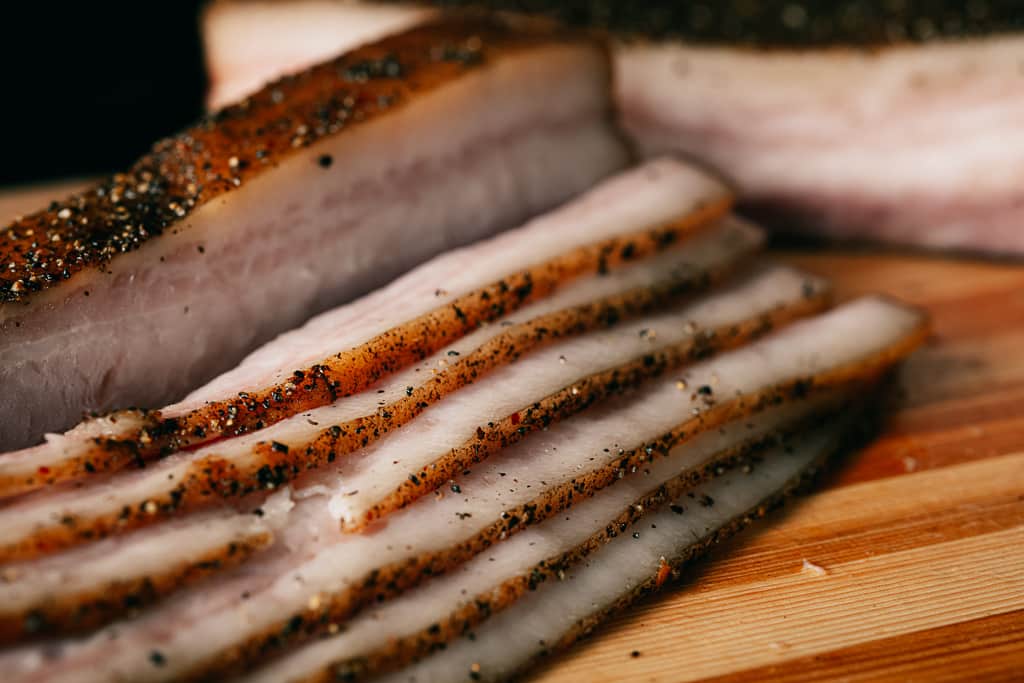 Sliced pork belly into bacon strips on a wooden cutting board.