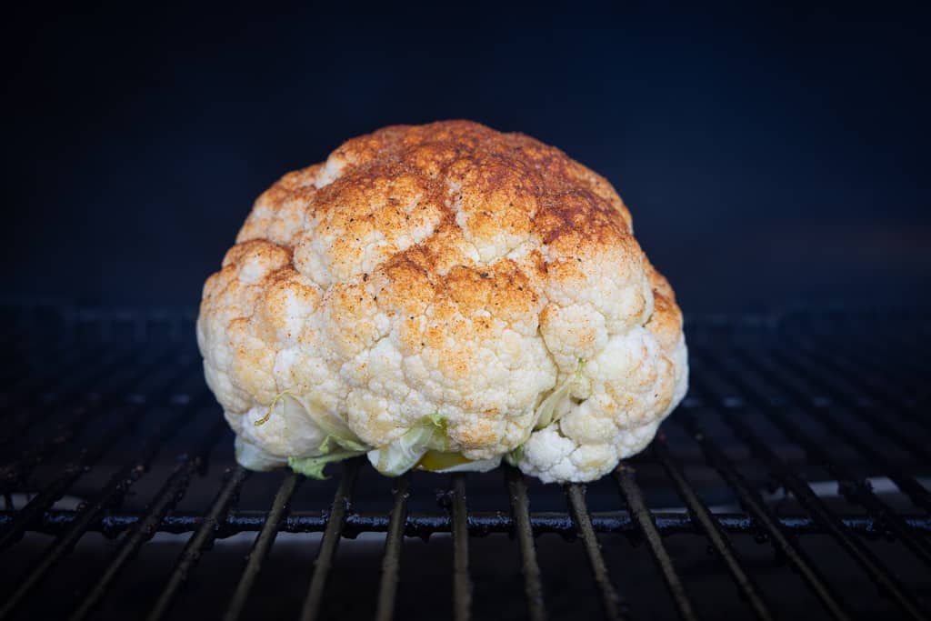 Seasoned whole cauliflower on the grill grates of a grill.