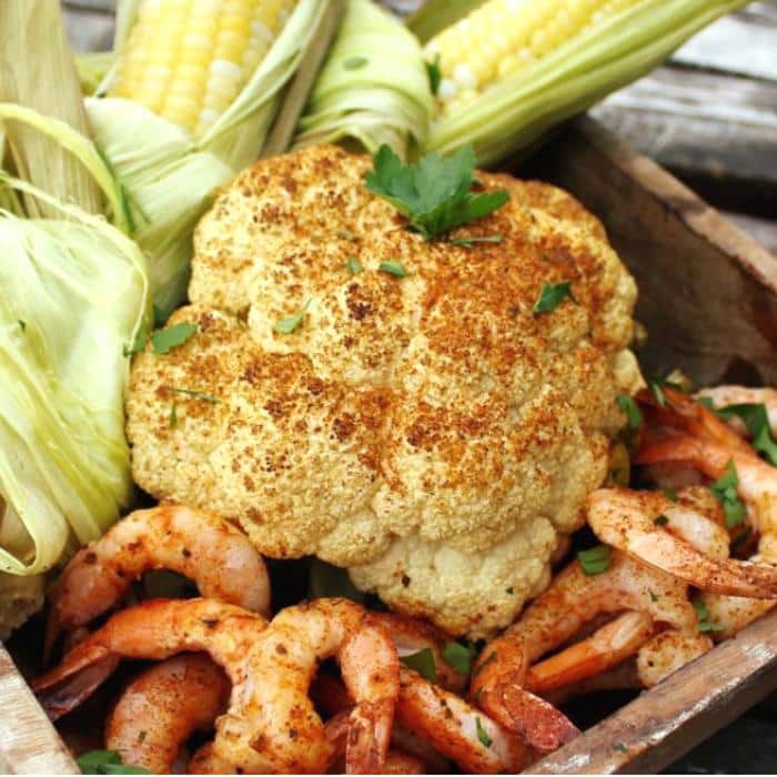whole grilled cauliflower next to grilled shrimp and corn