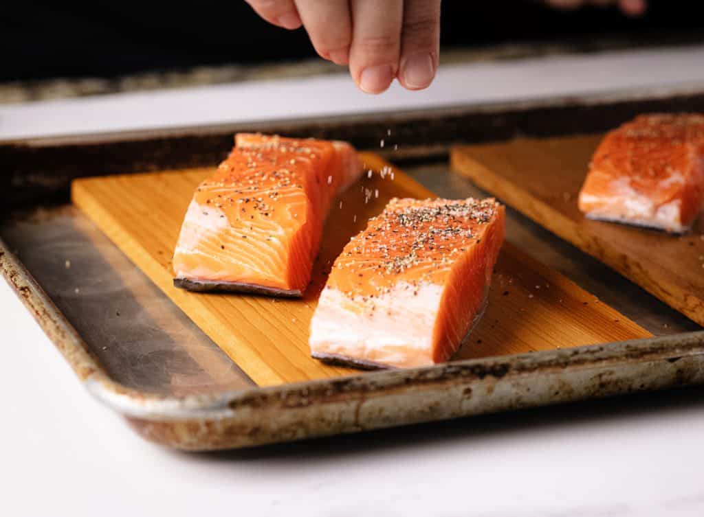 Salt and pepper being sprinkled on two salmon filets on cedar planks.