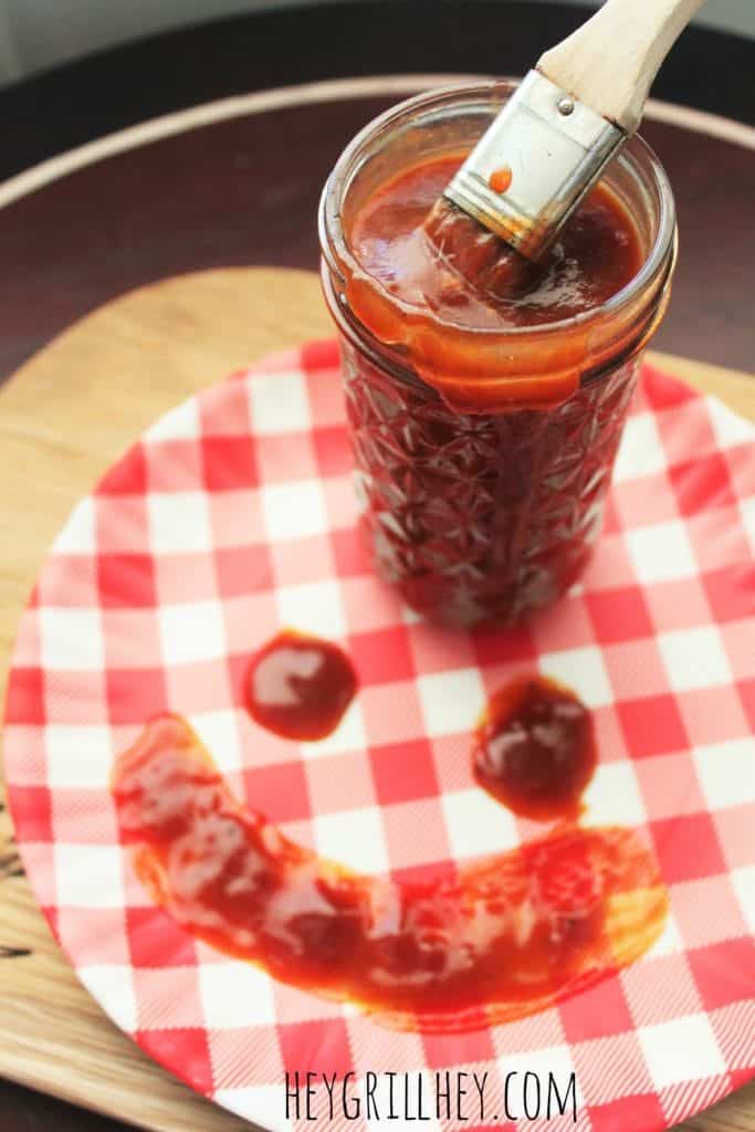 Jar filled with Barbeque sauce, resting on top of a red and white plate with a smiley face painted on it with BBQ sauce.