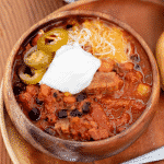 Wooden bowl of brisket chili topped with shredded cheese, sliced peppers, and a dollop of sour cream.