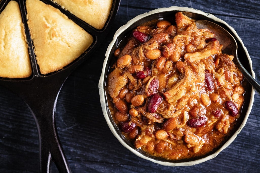 Bowl of pork belly bourbon baked beans in a bowl next to a skillet of corn bread.