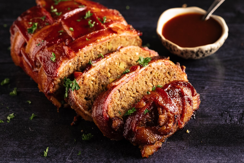 Sliced bacon weave wrapped meatloaf next to a bowl of BBQ sauce.