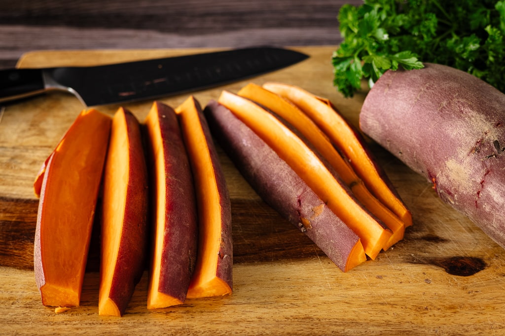 Sliced sweet potato spears on a wooden cutting board with a knife on the board in the background.