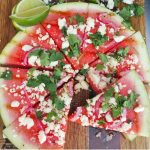 large circular slice of watermelon sliced into triangles as pizza shapes- grilled and topped with feta cheese and cilantro, served on a wooden cutting board