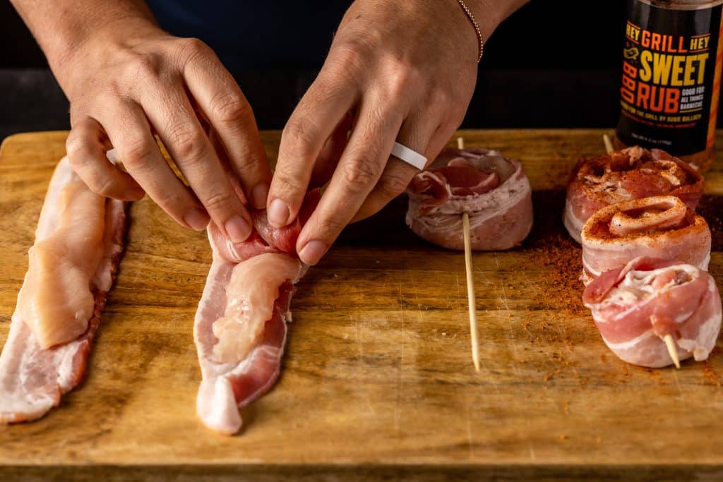 Chicken strips on bacon being rolled and skewered.