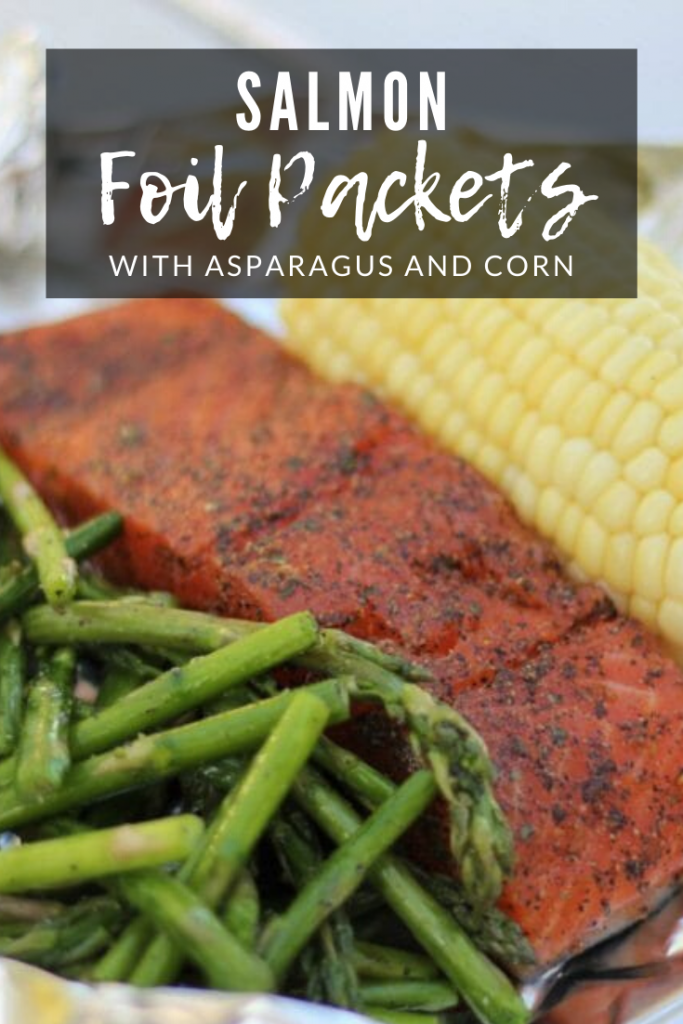 Grilled asparagus, seasoned whole salmon, and an ear of corn on a sheet of aluminum foil with the text overlay: "Salmon foil packets with asparagus and corn."