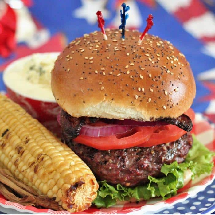 hamburger topped with lettuce, tomato, onion, bacon on a seeded bun, sitting on a plate next to a corn on the cob.