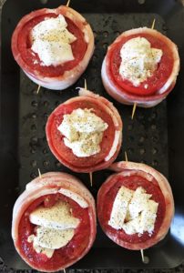 aerial view of five hollowed out uncooked tomatoes filled with mozzarella cheese and wrapped in bacon, with a toothpick holding the bacon around the tomato