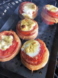 Five hollowed out tomatoes stuffed with mozzarella cheese, each wrapped in a slice of bacon, inside the grill on a grill grate surrounded by smoke