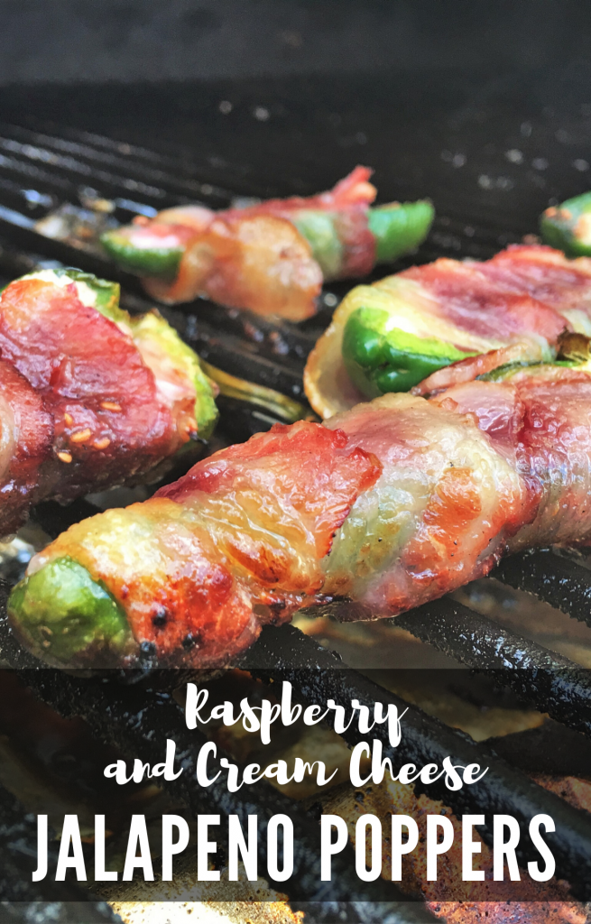 Jalapenos sliced in half, stuffed with cheese and wrapped in bacon, spread out on the grill grate. Text overlay reads "Raspberry and Cream Cheese Jalapeno Poppers."
