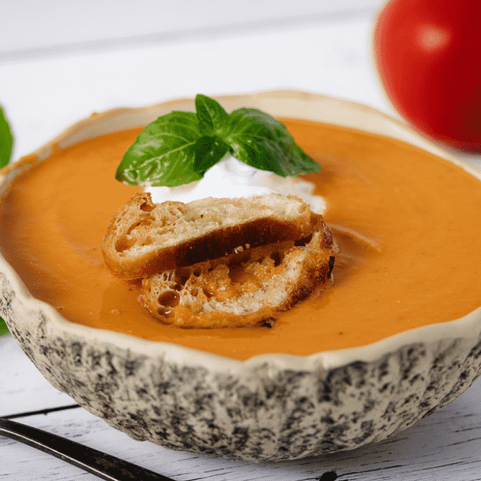 Bowl of smoked tomato soup next to a spoon, fresh basil, and fresh tomatoes.