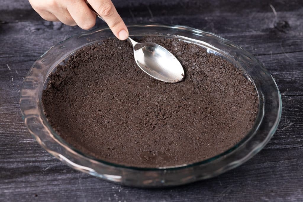 Cookie crust being formed into a pie dish with the back of a spoon.