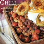 wooden bowl full of smoked chili topped with sour cream, cheddar cheese, and crispy jalapeno rings