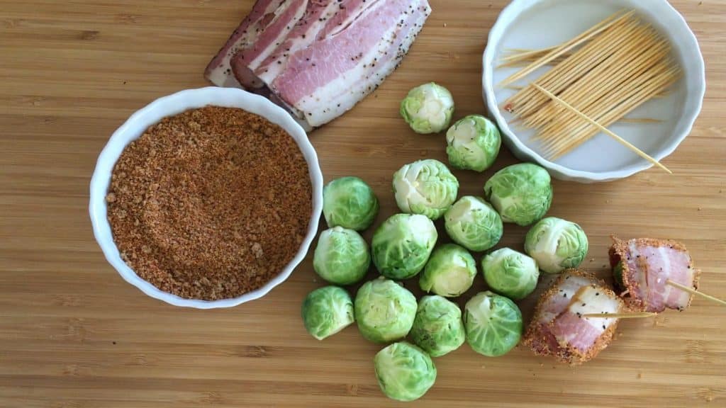 Whole brussels sprouts on a wooden cutting board surrounded by a bowl of BBQ rub, uncooked peppered bacon, long toothpicks soaking in a bowl of water, and two bacon wrapped brussels sprouts.