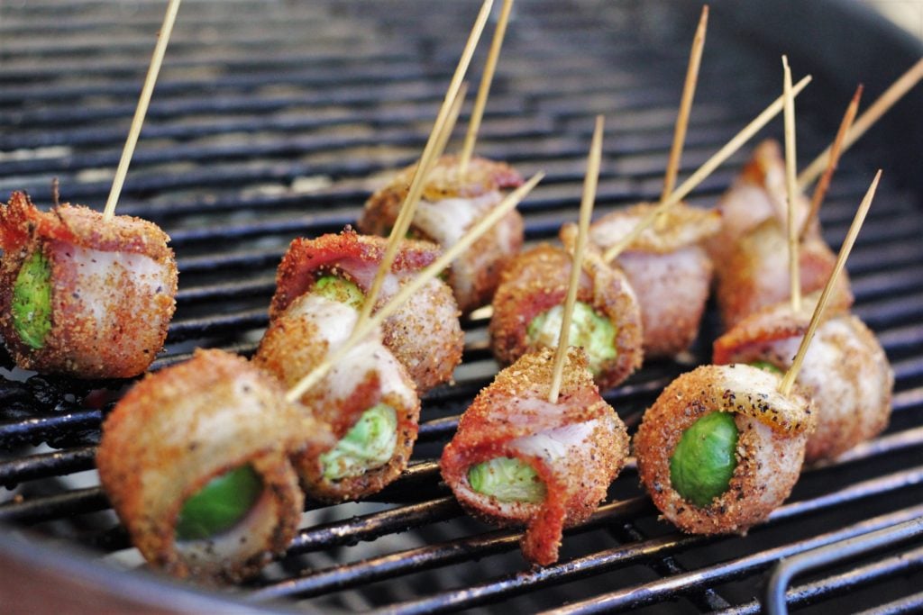 Bacon wrapped and seasoned brussels sprouts secured with long toothpicks on the grill grates of a charcoal grill.