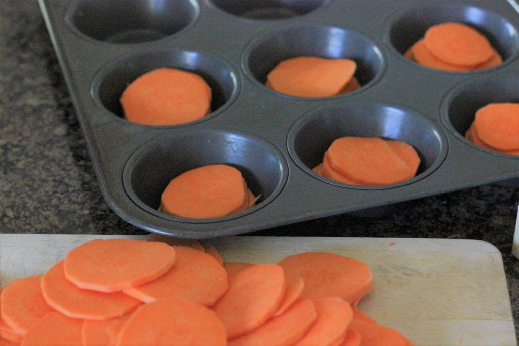 Uncooked sweet potato slices stacked in a muffin tin next to a cutting board of sliced sweet potatoes.