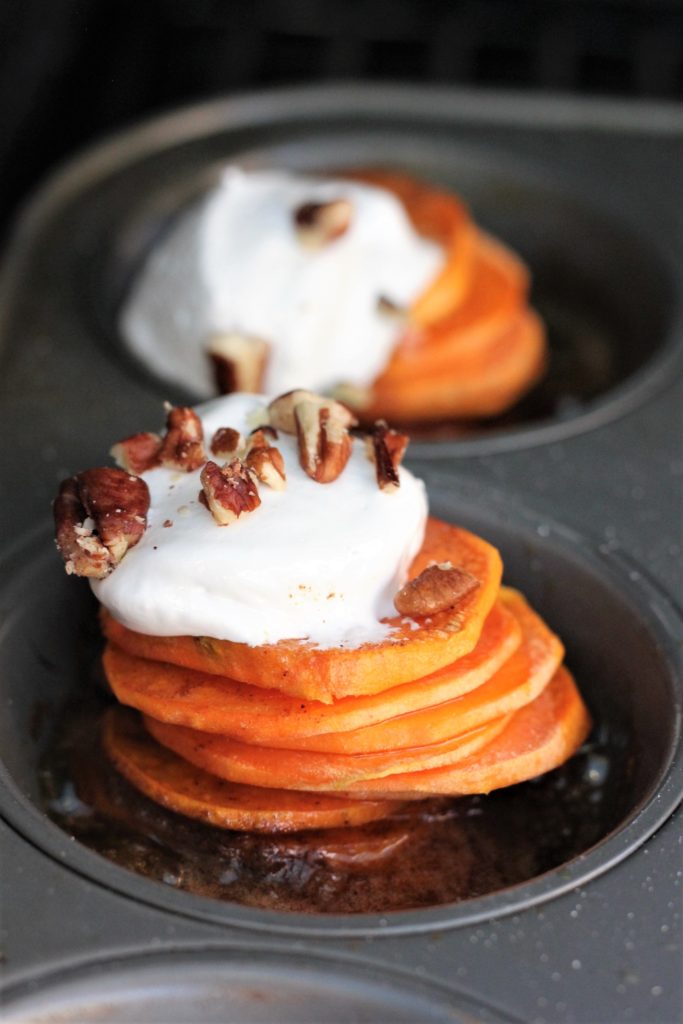 Two stacks of sliced sweet potatoes in a muffin tin, topped with a spoonful of marshmallow fluff and a half dozen chopped pecans.