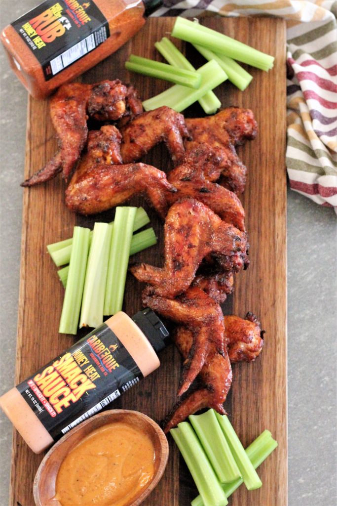 Aerial view of wings and celery arranged on a wood cutting board with bottle of rub and bottle and small wood bowl of sauce.