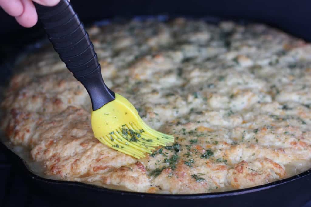 Yellow basting brush spreading garlic butter and parsley on top of the cheddar biscuit crust of a skillet pot pie.