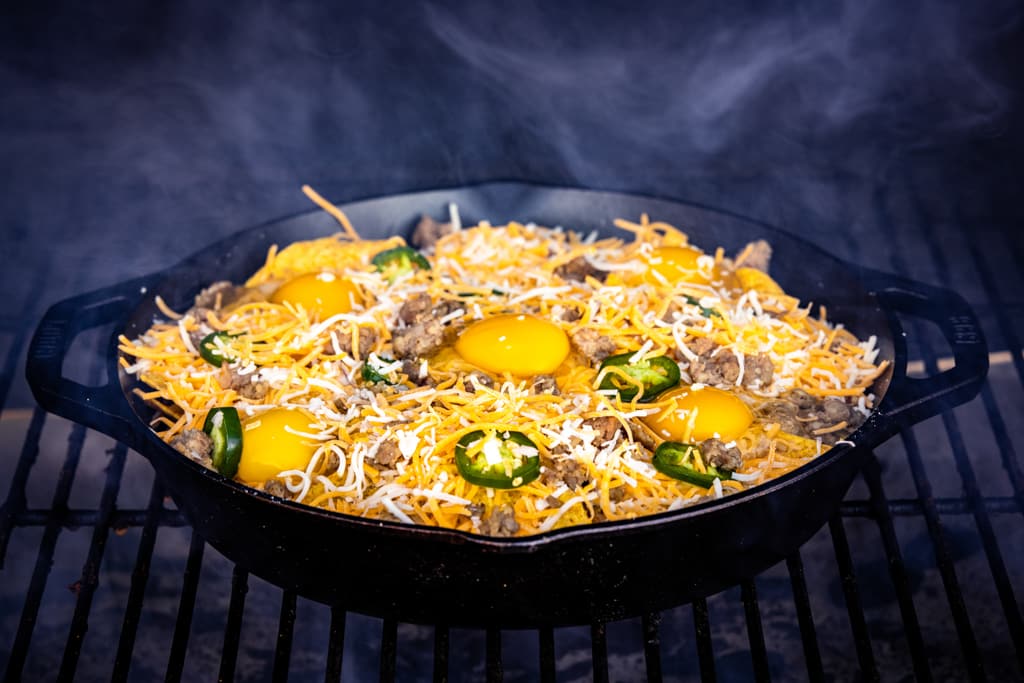 Loaded breakfast nachos in a cast iron skillet on the grill grates of a smoker surrounded by blue smoke.