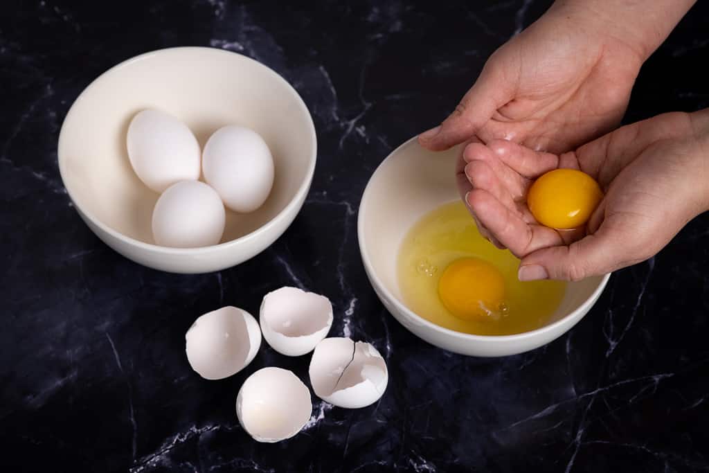 A pair of hands holding an egg yolk with a bowl containing one whole egg underneath next to a bowl of three whole eggs and two opened egg shells.