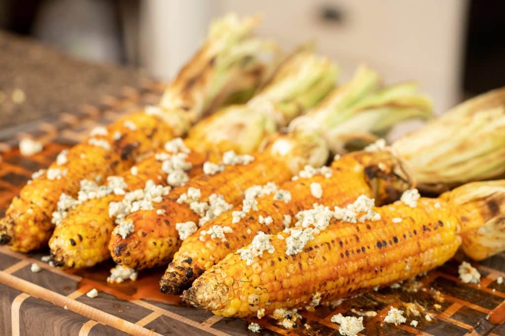5 ears of grilled corn on a wooden cutting board with blue cheese crumbled on top.