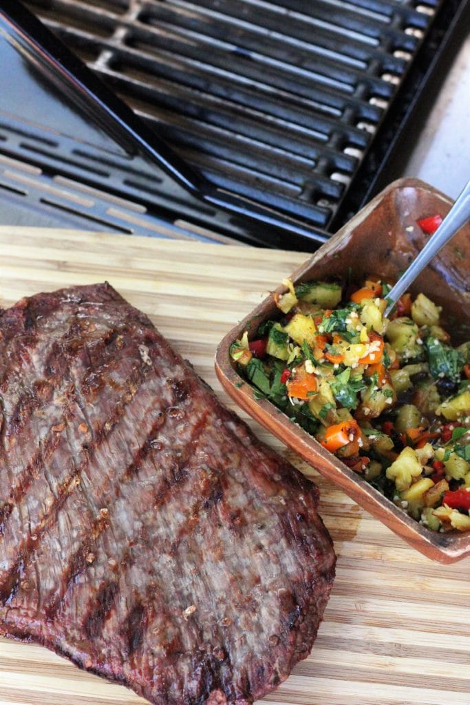 grilled salted flank steak on a wooden cutting board next to a bowl of grilled pineapple salsa