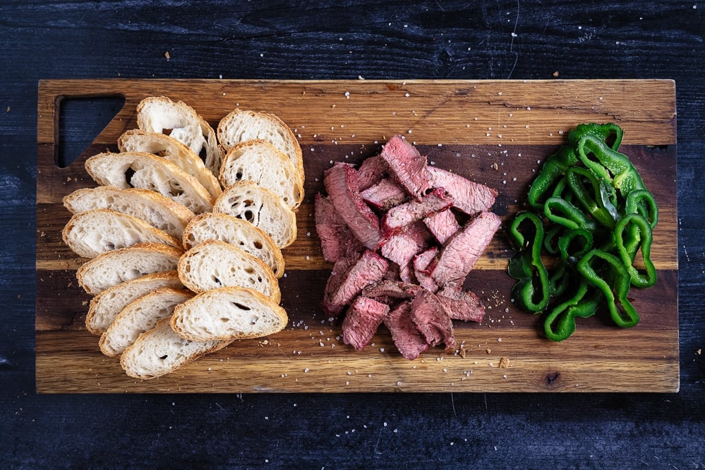 Crostinis, sliced steak, and sliced peppers on a cutting board.